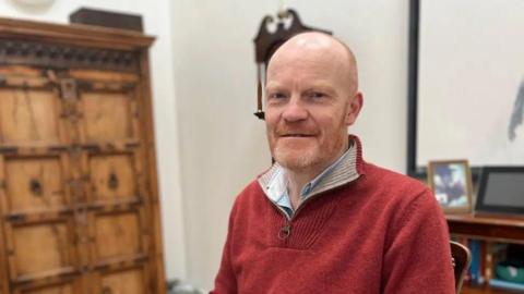 Deputy Gavin St Pier - A man with a red jumper, wearing a blue and white striped shirt with a ginger and grey beard.