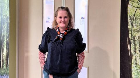 Jo Argyle is standing in front of a hospital door with her hands in her jeans pockets. She is smiling at the camera.