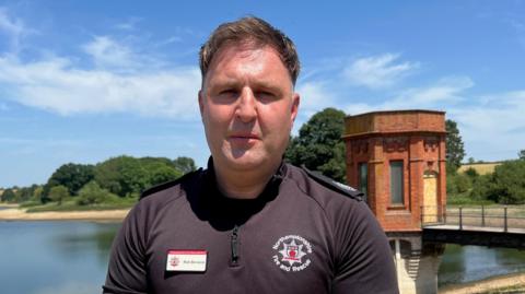 A man in a black Northamptonshire Fire and Rescue Service branded shirt with a name badge. He is posing against the backdrop of Sywell Country Park.