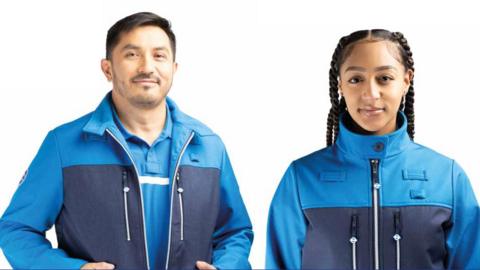 A man and a woman wearing TfL's new uniform, an outer layer where the top, above the chest is blue and below is a darker navy blue with two horizontal pockets next to the zip. 