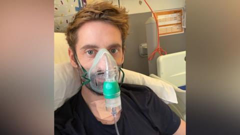 Daniel Harding wears an oxygen mask and looks into the camera. He has mid-length dark blond hair and is wearing a black T-shirt and is lying in a hospital bed. 