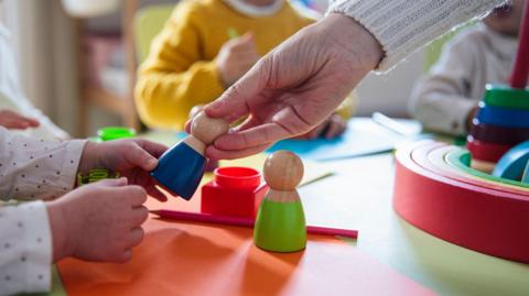 A close-up of an adult hand passing a wooden toy into a child's hand.