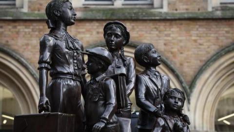 A life size bronze statue of five children in 1930s clothes, with a large brick building in the background