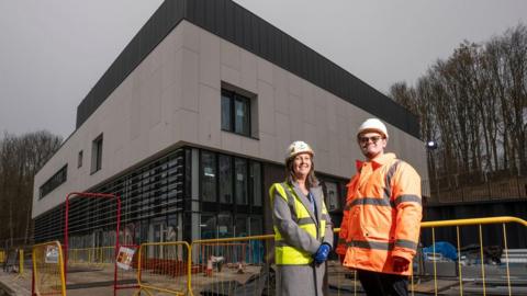 Durham County Council councillors Karen Allison and Joe Quinn standing outside the museum. They are both wearing hard hats in front of the grey, cube-shaped building. Allison is in a yellow hi-vis waist coat and Quinn has on an orange hi-vis jacket. Allison has shoulder length dark hair and Quinn has short hair and glasses. Both are smiling.