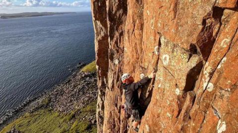 Alex Jones climbing a rockface in a coastal area. He is wearing a white helmet and climbing the red cliff, which gives onto a bay.