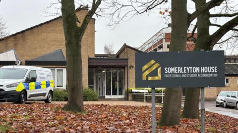 A white police forensics van is parked outside of an accommodation block on Somerleyton Street in Norwich. In the foreground of the picture is a grey sign which says SOMERLEYTON HOUSE, PROFESSIONAL AND STUDENT ACCOMMODATION.