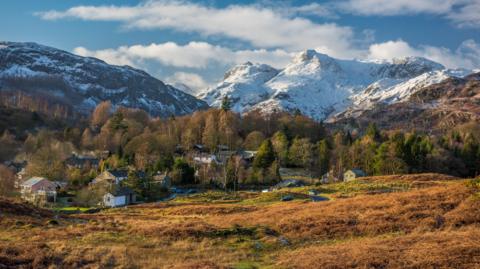A general view of Elterwater and Langdale Pike covered in snow. There are a few houses at the bottom of the fell, surrounded by trees and open green space.