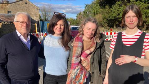 John, Sam, Harriet , Michelle stand together in front of the road closure on a sunny day. John has white hair and wears glasses, a black cardigan and a light-coloured shirt. Sam is wearing a blue and cream jumper, and has long dark hair. Harriet is wearing a khaki coat and a large scarf, and has a light-coloured bob, while Michelle is wearing a red striped t-shire and black dungarees. 