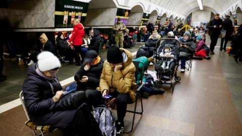 People sheltering inside a metro station, all dressed warmly. Most are sitting on fold-out chairs, while others are on the floor or standing. There is also a baby in a pram.