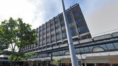 A five-storey office block viewed from an angle slightly to the side rising above a shopping centre. It has brown bricks and rectangular windows that are all the same size.