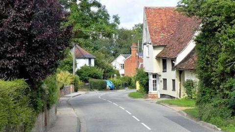 The Street in Monks Eleigh. The road is lined by small cottages, tall trees and leafy bushes. It is a 30 miles per hour limit and is slightly bendy.