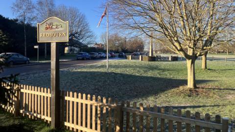A picture taken in Redbourn village in Hertfordshire. There is a sign that says Redbourn at the front of the picture, in the middle a union flag at half mast plus a war memorial. On the right of the image there are two trees and the sky is blue in the background.
