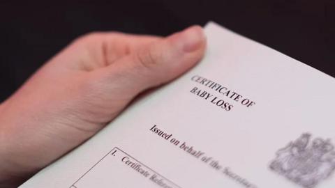 A hand holds a the corner of a white baby loss certificate, which was issued in England. 