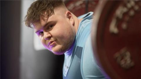 Archie Cleverley looking sideways at the camera while standing with a barbell behind his shoulders with red weights on either end. He looks to his left at the camera. He wears a blue top.