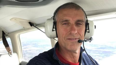 A smiling middle-aged man in the seat of a small plane, flying above fields. He is earing a headset.