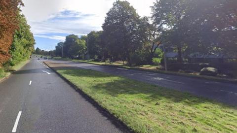 The H10 Bletcham Way road as it turns from dual to single carriageway. There is a grass verge in the middle and autumn-coloured trees on the sides of the road. No cars are pictured on the road, except in the distance.