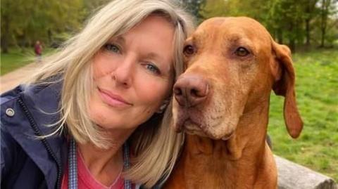 Sam Carter is wearing a red t-shirt and navy blue jacket sitting next to her dog in a park