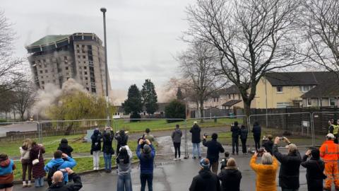 People gathered behind a metal fence taking photos and videos of a building being demolished. The building is in the background and is seen collapsing.