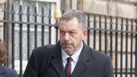 Tony Chambers, who has short grey hair and a grey beard, walks towards Liverpool Town Hall wearing a dark grey suit and sets his face in a serious expression