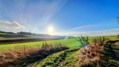 The sun rises in a blue sky over green fields in the foreground 