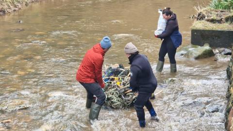 This picture shows volunteers from British Divers Marine Life Rescue clearing plastic waste from Boggle Hole, near Robin Hood's Bay. Two volunteers, stand ankle-deep in the water dragging a large bundle of litter. A third volunteer stands behind them, holding a bag of waste.