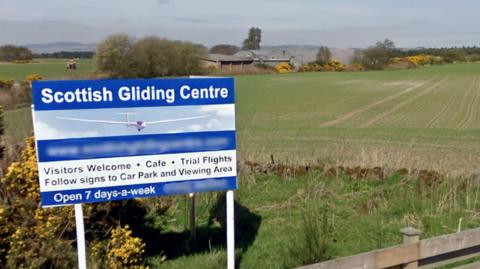 A rural road runs alongside open fields under a clear blue sky, with a sign for the Scottish Gliding Centre visible near a wooden fence.