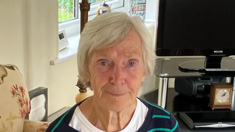 Veronica Bird. She has short blonde hair and sits in a living room. She is wearing a white T shirt and stripey jumper.