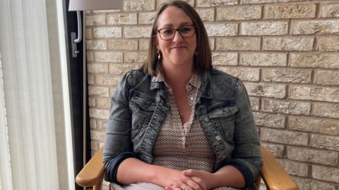 Nicola Stanyon smiles, sitting in a wooden chair with her hands clasped together, wearing glasses and a blue denim jacket with pink patterned top