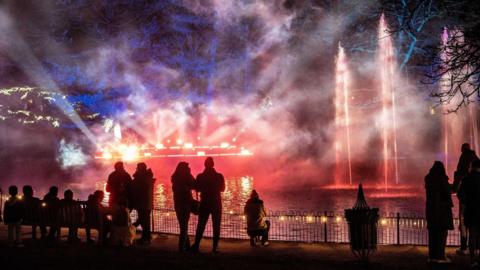 A pond in a park is filled with smoke and purple and red lights. There are fountains of water spraying into the air. People are standing in the park watching the display.