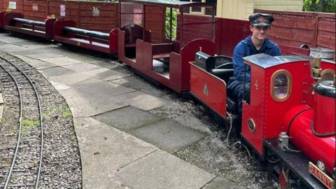 A train driver, who is wearing blue overalls and a black cap, is sitting on the miniature red train. Behind him are three carriages for visitors to enjoy the ride.