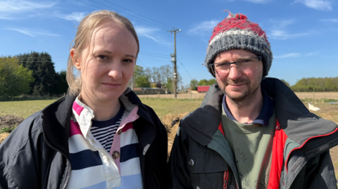 A woman and a man stand in front of a green field under a blue sky. The woman has blond hair tied back and is wearing a rugby shirt with blue, white and pink stripes and a black jacket. The man is wearing a grey and red bobble hat, glasses, a grey coat and a green jumper.
