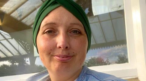 A woman wearing a green headwrap and denim jacket sits outdoors in front of a window.
