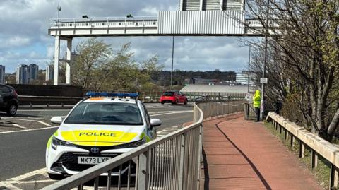 Front view of a police vehicle parked at the side of a road at the end of a bridge. Other cars can be seen on the road and a figure in a high viz jacket stands next to a section marked off by police tape.