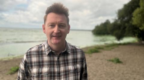 Neil Reid, who has spiky brown hair and a checked shirt stands in front of a green lough