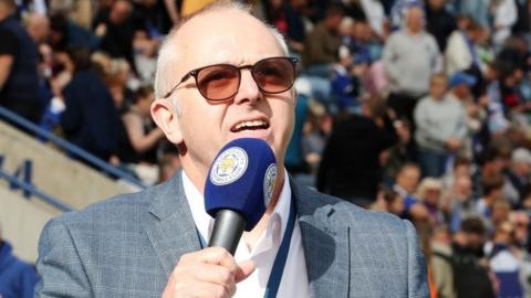 Bradley Varnam speaking into a microphone in front of full stadium seating