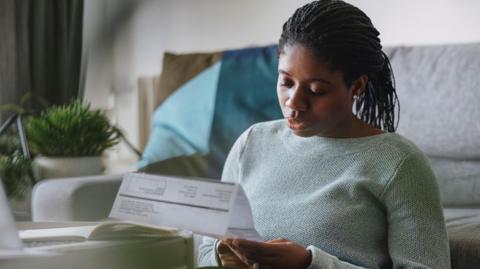 A person looking at a bill as she sits in front of a sofa