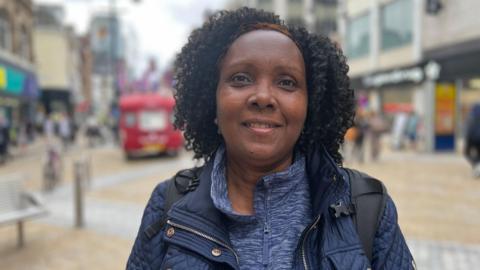 A woman with black curly hair looks at the camera. She wears a blue padded jacket and blue roll neck jumper. Backpack handles are strapped across both of her shoulders. Behind her, Briggate is blurred.