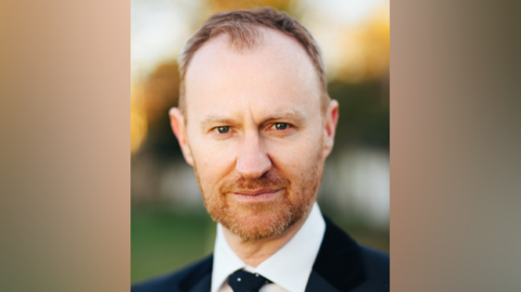 Mark Gatiss is looking at the camera and wearing a blue suit. He has short brown hair and a ginger beard.
