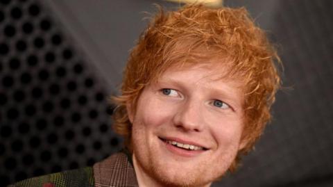 Ed Sheeran looks away from the camera and is smiling. He has red hair that covers his forehead and ears.