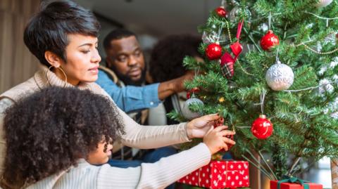 A woman decorating her christmas tree. Her family surround her. There are red and silver baubles on the tree. 