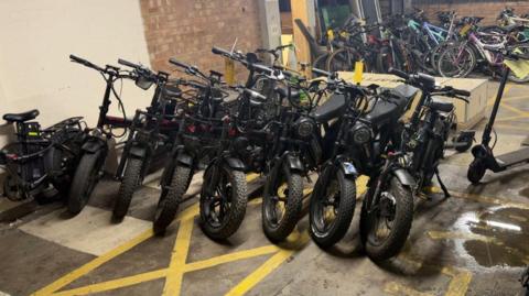 A line of illegal black e-bikes with multiple other bicycles in the background, in a police storage area.