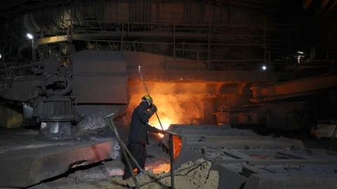 A worker in a hard hat stokes the furnace, which is bright orange. There are steel objects around the man.
