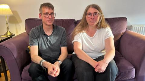 Chris, a man with glasses and wearing a grey polo shirt, is sat on a purple sofa next to his wife Diana, who had long light brown hair and is wearing glasses and a white T-shirt. They are both looking at the camera