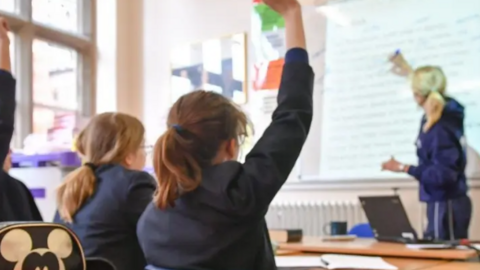 Two schoolchildren pictured in a classroom. The girl on the right has raised her hand and a teacher is pointing to something on a whiteboard. The teacher has blonde hair and is wearing a navy hoodie and joggers. The girls are wearing navy blazers. 