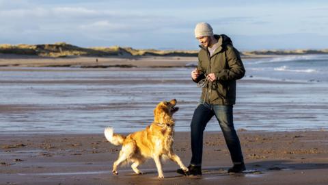 A young man and his golden retriever walk along a beach. 