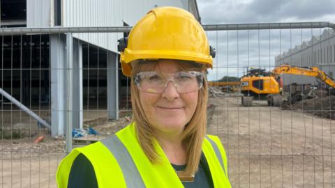 Sarah Jones is wearing a yellow hard hat, a high-vis vest and protective glasses while standing on the construction site at the Harland and Wolff shipyard. She is stood in front of a metal fence, and behind her are yellow diggers and an unfinished tin warehouse.