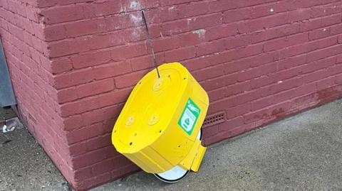 A yellow defibrillator which has been kicked off the wall and as hanging by a wire.