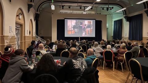 People sit around round tables on black chairs with gold trim. They look up at a large screen showing a film.