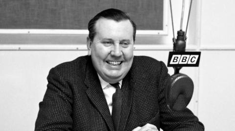 A black and white picture of Malcolm Arnold sat behind a 91ȱ microphone, smiling and wearing a suit and dark tie.