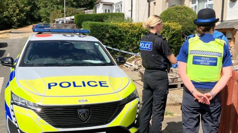 A female police officer is standing next to a police community support officer in a street. They are next to a police car and facing a row of houses.